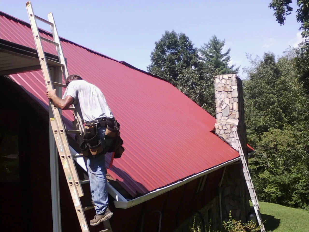 Roofer on a ladder beside a red metal roof and stone chimney — Soffit & Fascia Repair by Roofing Brothers in Bell
